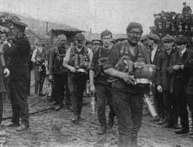 A team of 6 or seven men, covered in coal dust and each carrying their respirator packs. They are watched by a crowd of onlookers.