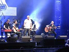 A stage shot with four men clearly visible. The man at left is leaning at a rectangular box, he has a microphone nearby. The second man is shown in right profile, he is playing a guitar and singing into a microphone while seated. The third man is standing in the middle of the stage with a arms at his sides, he carries a microphone in his right hand. The fourth man is also seated while playing a guitar and at a microphone. A fifth man is obscured seated behind and to the right of the middle one. The men are surrounded by stage equipment including speakers, lights and additional microphones. A drum kit is partly visible but the drummer is obscured.