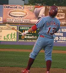 A man in a light-blue baseball uniform with maroon trim reading "Harman" and "13" on the back; he is holding a baseball bat in his left had and wearing a maroon baseball helmet atop his head