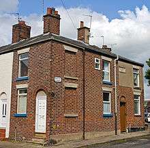 A two-story brick terraced house with chimney pots on its roof, attached to its neighbour, under a blue sky with clouds. Two of the windows on the front have been bricked in, and a small portion of the facade at upper right is concrete. A small black and white plate on the front identifies it as being on Barton Street. Around the corner is a white wooden door in a brick arch; at right is a similar doorway with a brown door.