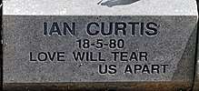 A gray stone with "Ian Curtis, 18-5-80, Love Will Tear Us Apart" carved into it in block letters