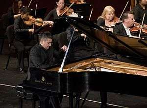 A man plays a piano with enthusiasm, his left hand raised in preparation for striking keys; behind him, the string section of an orchestra plays accompaniment.
