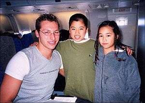A young man is at left wearing a tight grey T-shirt with white sleeves, with "French Connection" across his chest. He has short hair and is wearing elliptical glasses. In the centre is a boy with very short black hair in a green T-shirt, and to the right is a young girl with pigtails wearing a blue coat. They are posing on an aeroplane.