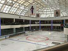 An ice hockey rink seen from a second story above it. There are two teams playing at the far end. People are watching the game from both levels; there are stores behind them. Above the rink is a glass ceiling from which advertising banners hang promoting the tournament sponsors, as well as the Canadian and U.S. flags