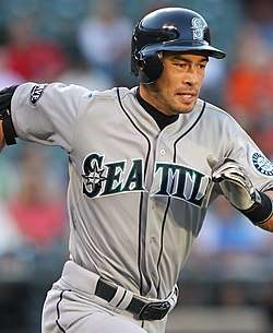 A man in a gray baseball uniform running to the right. The uniform reads "Seattle" across the front, and has a sleeve patch with a teal and silver compass surrounded by "Seattle Mariners". The man is wearing a dark batting helmet.
