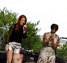 Two women singing; one is wearing a long-sleeved black shirt and white shorts and the other one is wearing a dress.