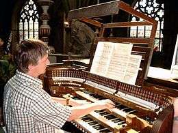 an organist seen from the right at his organ with three manuals, gothic church windows in the background