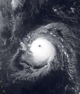 Satellite image of a slightly elongated tropical cyclone over blue waters; an eye, visible as a void at the center of the mass of white clouds, is visible. Small green islands dot the lower-left corner of the image.