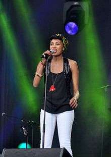 Shot of Imany performing in front of a green backdrop, wearing a black top along with white pants.