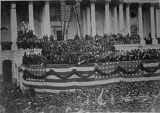 Photograph of a crowd in front of Capitol building decorated with patriotic bunting