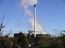 The tops of a row of trees at the foreground obscure a large industrial building. In the centre, reaching to the top, is a white thin chimney and behind this trails white smoke into the sky.
