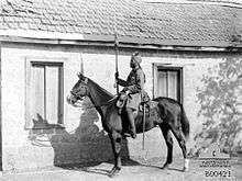 In a black and white photograph, a man wearing a turban and military uniform sits astride a stationary, dark-coloured horse, facing left. In his right hand he holds aloft a sword. Directly behind them stands a single-storey, brick building with two large windows and a tiled roof.