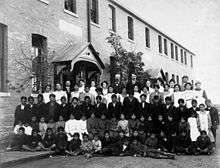Posed, group photo of students and teachers, dressed in black and white, outside a brick building in Regina, Saskatchewan