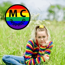 An image of a young woman wearing a rainbow-colored sweater and jeans. Her hair is pulled up in a ponytail with flowers. She is sitting in grassy field. There is a circle filled in by horizontal rainbow stripes. The letters "M C" and "Inspired" are included in the circle in a black font.