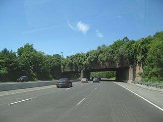 A six-lane freeway in a wooded area with an overpass containing trees