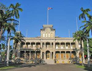 Photograph of the front of &#x02BB;Iolani Palace, flanked by palm trees with the Hawaiian flag flying atop the central tower.