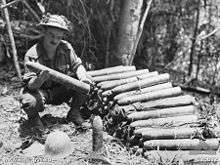 A soldier kneels beside a pile of artillery shells in a clearing in the jungle, closely inspecting one that he is holding