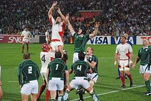 Two rows of opposing players, green to the fore, white behind, each aid a jumping player from their team by lifting him towards an off-picture ball travelling overhead