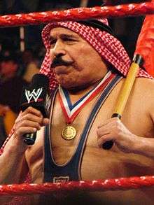 Behind two red ropes stands a man with a large mustache speaking into a microphone and holding a flag.