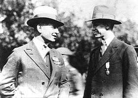 A mid-length black and white photograph of two men in suits engaged in conversation. They are outside, and have medals on their left breasts.