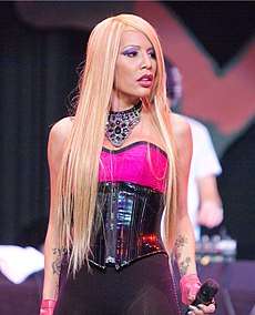A blonde woman looking to her left, wearing a pink and black&ndash;coloured dress and holding a microphone in her left hand.