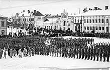 A parade of Finnish Jägers at the Vaasa town square. Spectators are gathered around the soldiers in the background. General Mannerheim is inspecting the formation in the foreground.