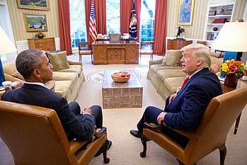 Barack Obama sits in the left Foreground while Donald Trump sits to the right with the ornate Resolute Desk center in the background.