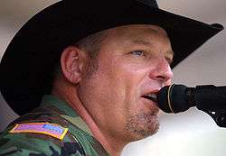 A man in a black cowboy hat and camouflage jacket singing into a microphone