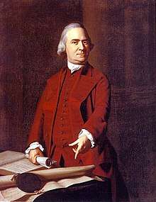 A stern middle-aged man with gray hair is wearing a dark red suit. He is standing behind a table, holding a rolled up document in one hand, and pointing with the other hand to a large document on the table.