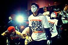 Four members of the hip-hop dance crew JabbaWockeeZ performing in a night club wearing white masks and white gloves.