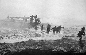 Black-and-white photograph of soldiers in uniform scrambling out of a Eureka boat into choppy waters while carrying ladders, with Churchill leading in the far-right, his sword visible in his right hand