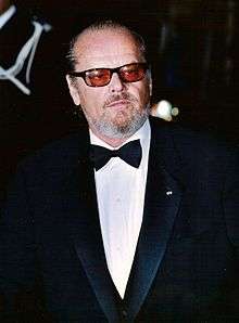 Photo of Jack Nicholson—a white man of 65 years, with gray hair and beard, wearing a black suit with a black ballot tie and wearing sunglasses with orange lenses—attending the Cannes Film Festival in 2002.