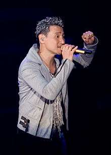 A man in a jacket with dyed hair singing into a hand held microphone