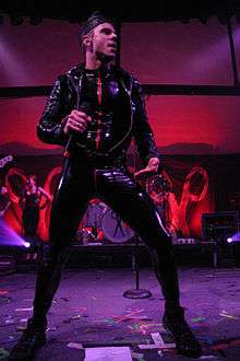 A Caucasian man wearing leather suit, performing on stage, facing right.