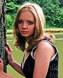 A Caucasian woman with blonde shoulder-length hair wearing a blue tank top with white trim looks to her left directly at the camera with her hands placed on a tree at the left of the image. Behind her is a body of water and some woods.