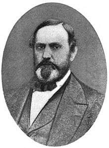 A man with receding black hair, sideburns, a mustache, and a goatee wearing a black jacket, vest, and bowtie, and a white shirt
