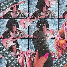 A woman's hand with pink fingernails holds a black television remote in front of duplicates of electric guitars and a picture of James Ferraro with sunglasses and lipstick. On the far right is a woman's hair.