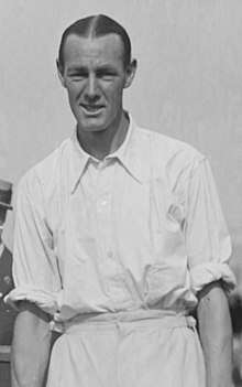 A black-haired man in a white shirt with sleeves rolled up looks into the camera, posing