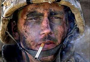 A man with dirt and ash on his face in a military helmet, staring intently forward while he smokes a cigarette