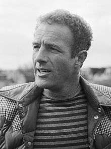 A black and white photo of a man in a striped shirt and jacket who has curly hair.
