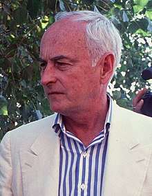 An elderly Caucasian man with white hair wearing a white jacket and a white-and-blue-striped shirt.