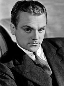 Black-and-white photo of James Cagney in 1930—a white man with serious features and an arched eyebrow, dark eyes and hair combed back, wearing a suit and around 30 years of age.
