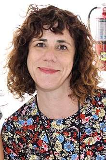 Headshot of Attenberg smiling at the camera. She wears curly hair shoulder-length with bangs and a floral-print top.