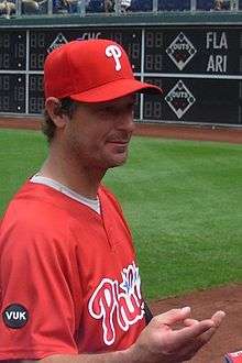 A man wearing a red baseball jersey and cap holds out his right hand