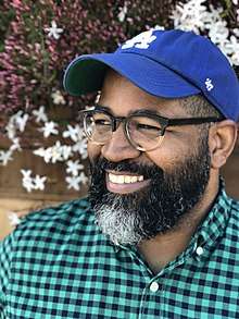 Headshot of Jamil Smith. He wears glasses, a green-and-black checked collared shirt and a blue Los Angeles Dodgers cap. He smiles, looking off-camera.