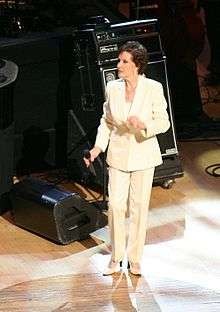 A dark-haired woman wearing a white jacket and pants, standing on a stage