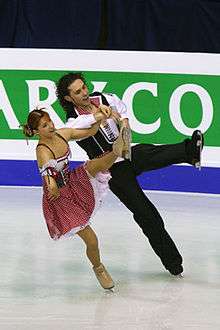 A man and woman ice dancing; the woman, on the left, has red hair pulled back tightly and is wearing a dark pink and white peasant dress, and the man is wearing a black and white shirt with a dark pink lapels and black trousers