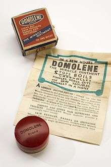 A jar of ointment, with a box and a poster. The box has the words "Domolene Brand Stops all skin troubles rashes and irritation The miracle ointment"