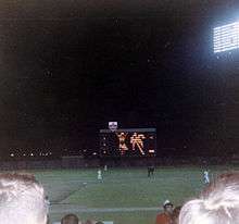 Fans watch a game from the third base line; the scoreboard is visible beyond the right field wall.