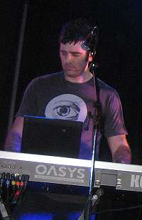 Upper body shot of a 34-year-old man. He is partly obscured by a keyboard and microphone in front of him. He has short dark hair and wears a dark tee-shirt, which has a large open eye logo. His eyes are directed down and slightly to his right. His arms are raised to the keyboard, but his hands are obscured. The lettering O-A-S-Y-S is visible on the back of the synthesiser with an additional K at the right.